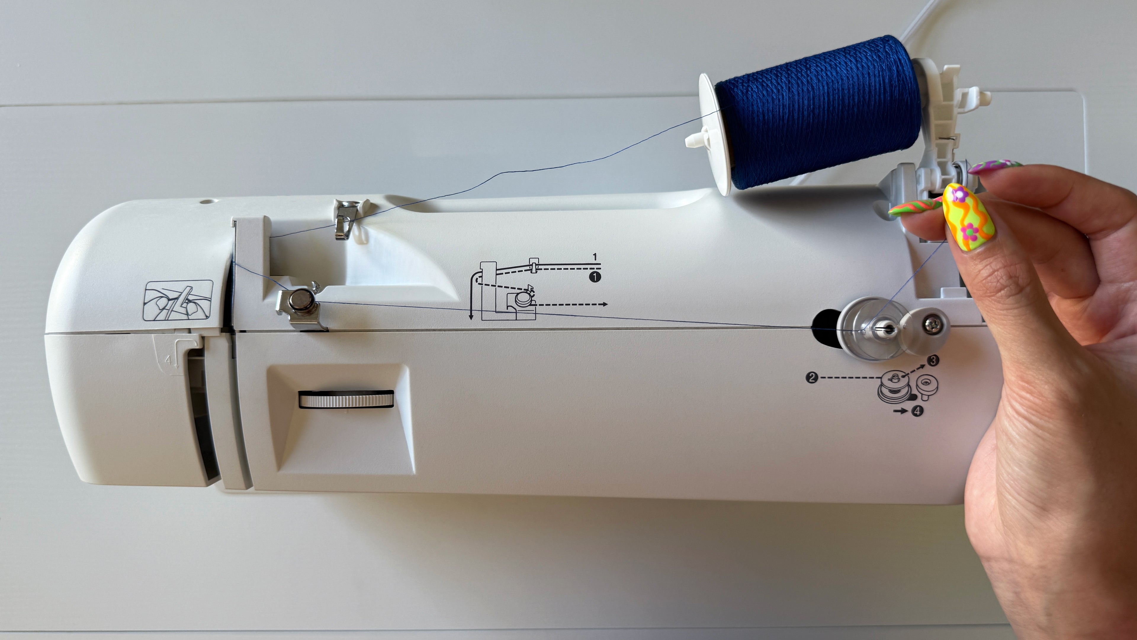 Sewing machine with a spool of blue thread and a hand holding the thread threaded through the bobbin