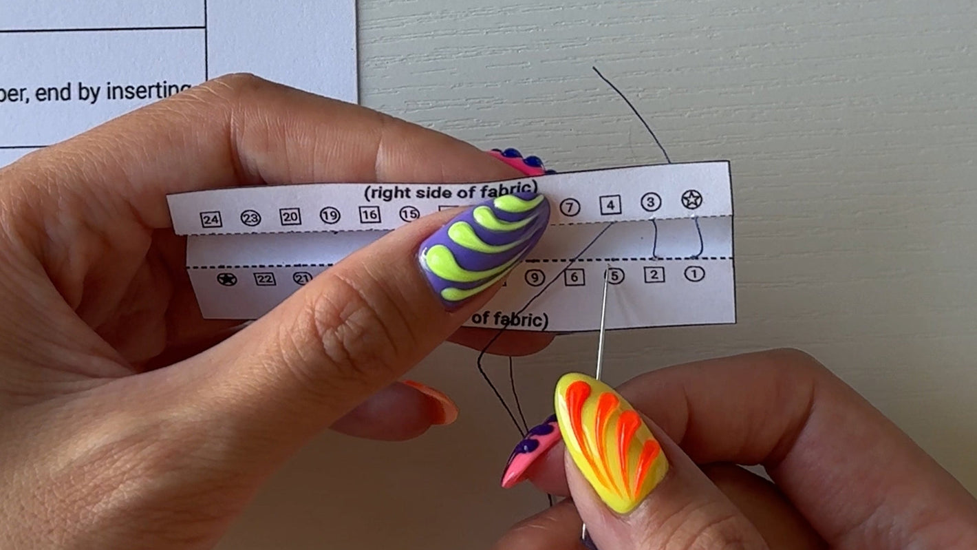 Close-up of hands with colorful nail polish holding a needle and thread, with a paper in the background.